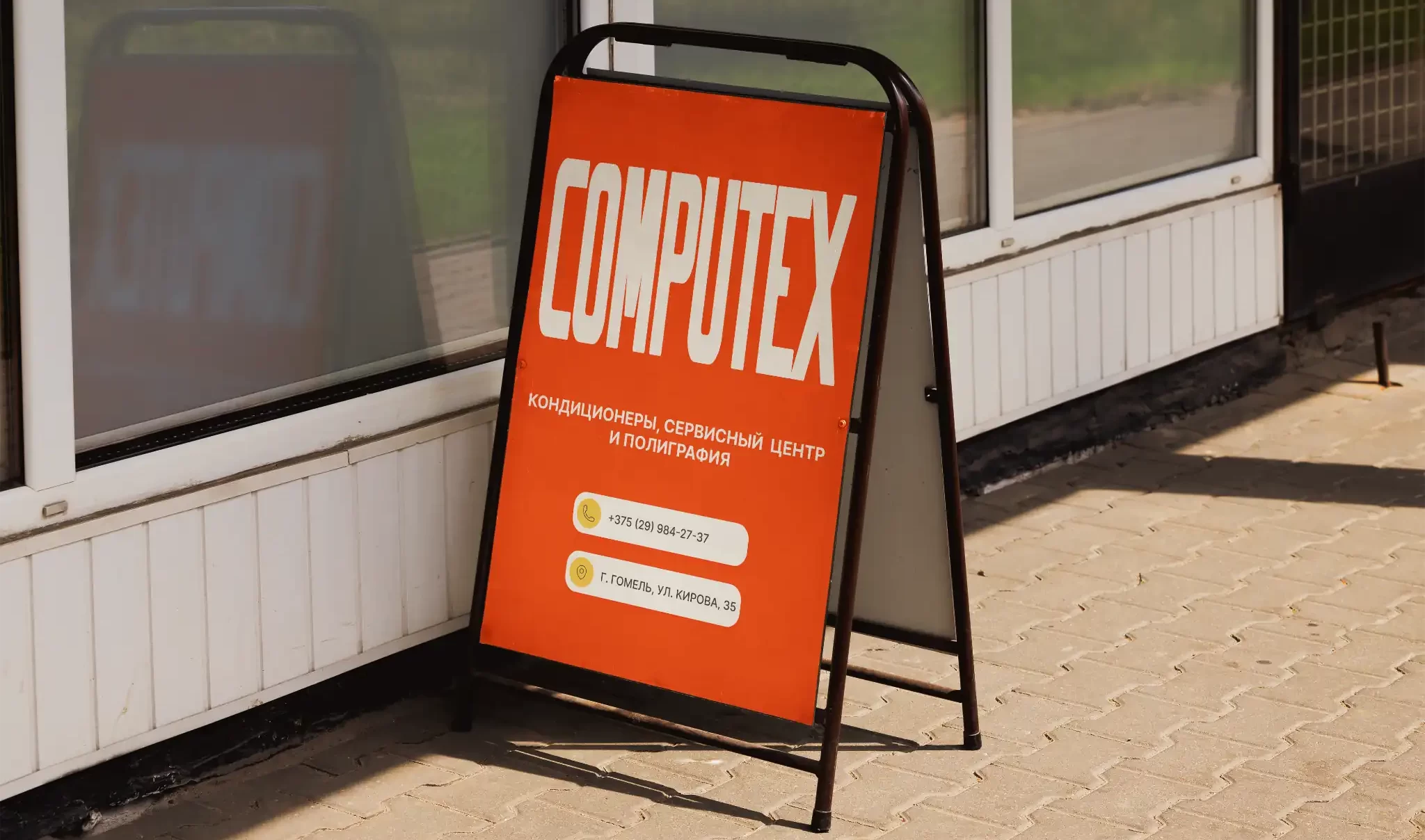Уличный штендер COMPUTEX с контактной информацией, установленный на тротуаре у входа в здание, с ярко-оранжевым фоном и белым логотипом